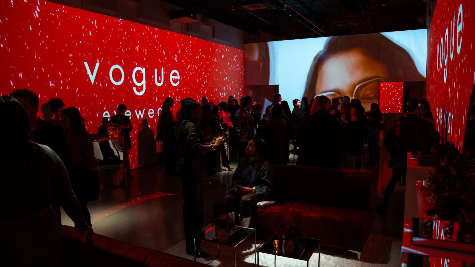 Guests mingling in a Vogue Eyewear launch space lit entirely in red, with a large screen showing a close view of eyeglasses and Vogue Eyewear branding projected across the walls
