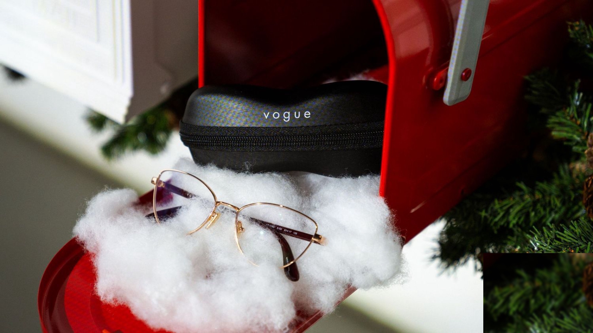 Vogue Eyewear eyeglasses with gold frames and a black Vogue Eyewear case resting on white cotton batting inside a red mailbox decorated with pine branches