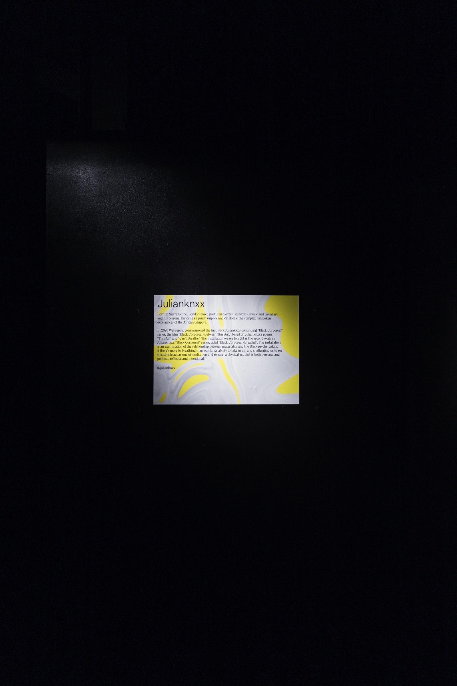 Small white wall text panel with yellow marbling lit against an otherwise black wall