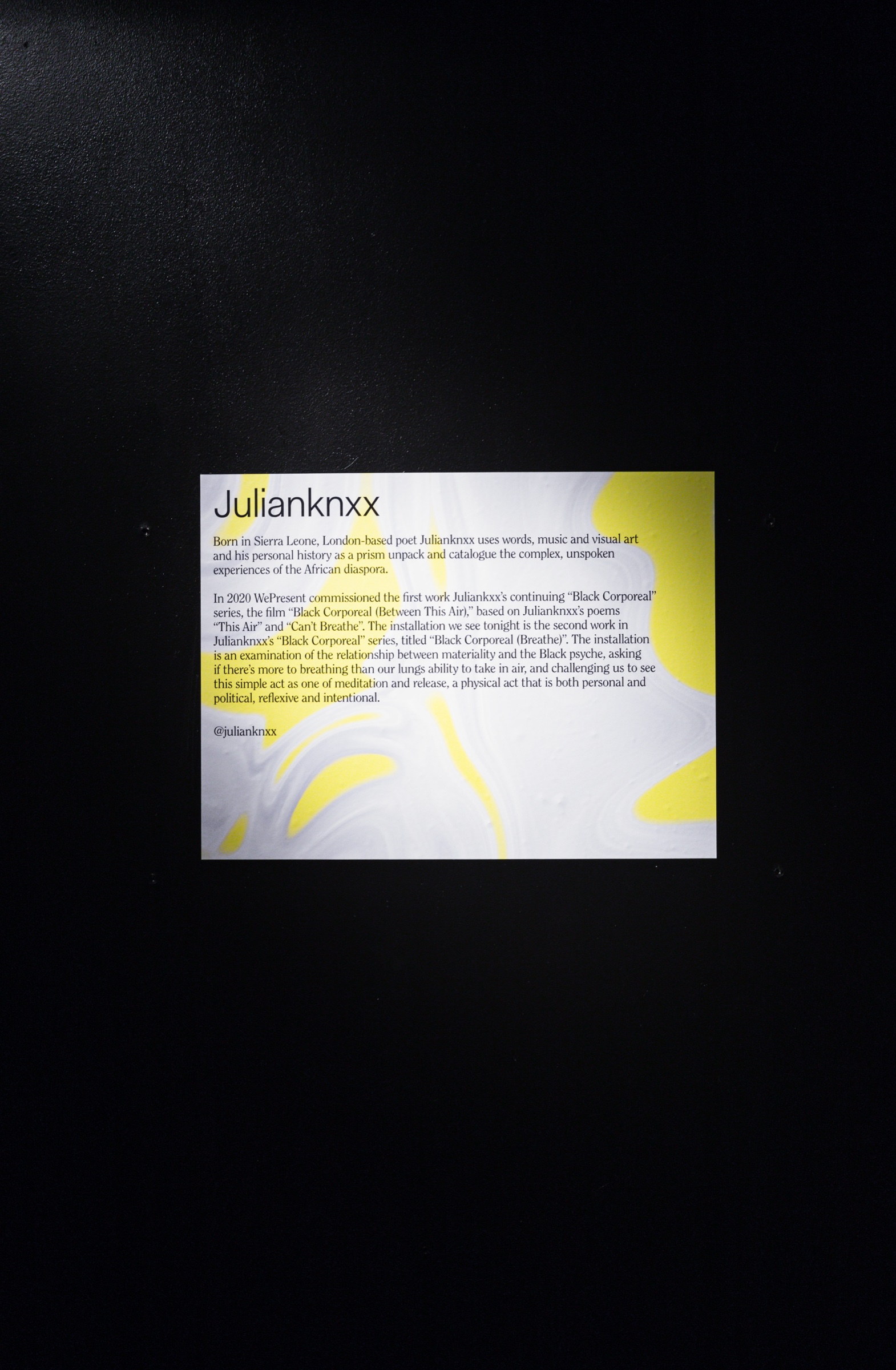 Wall label with a yellow splash describing the artist Julianknxx, mounted on a black wall