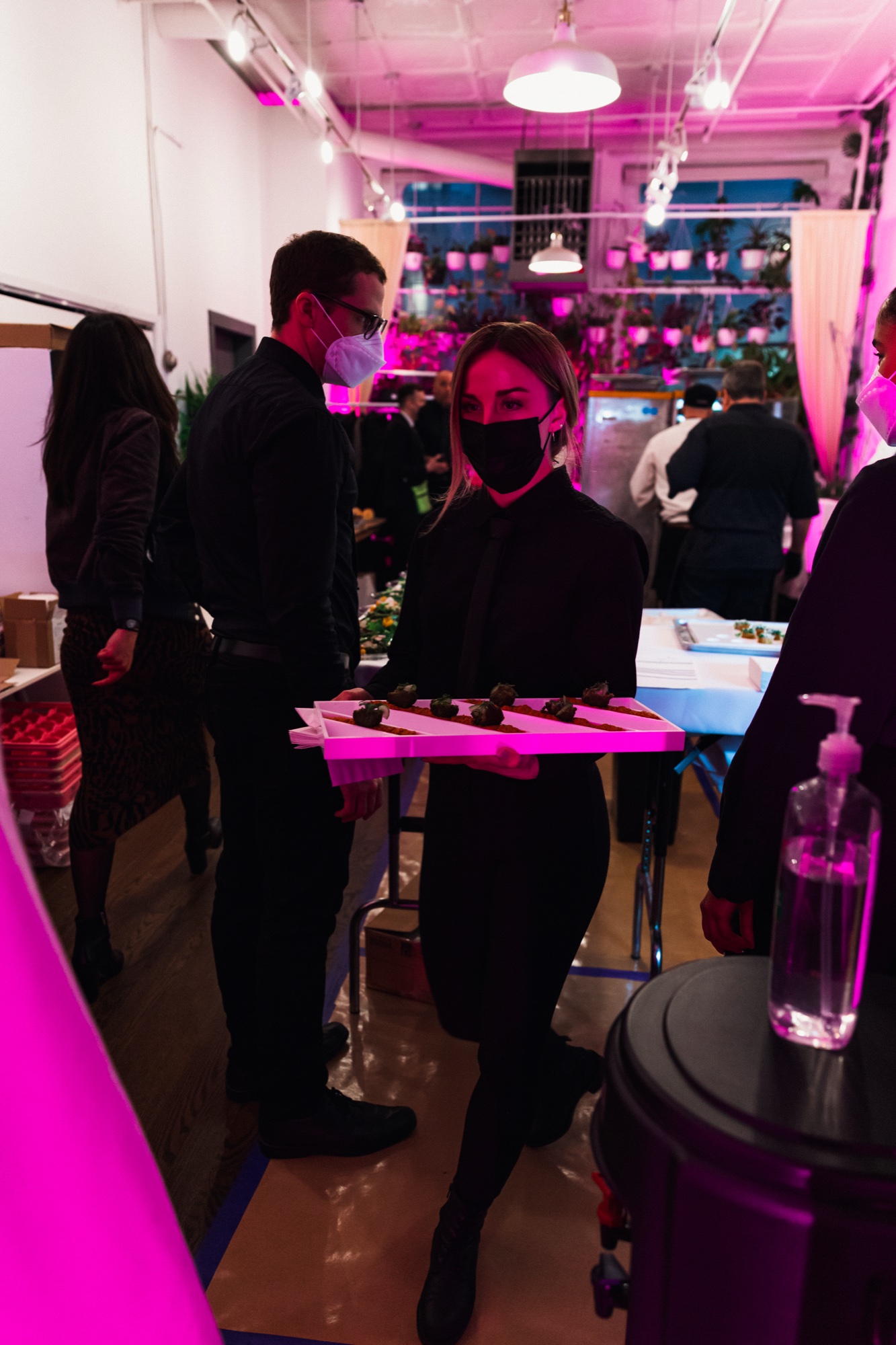 Masked server in black carries a pink tray of canapés through a magenta lit room with hanging plants
