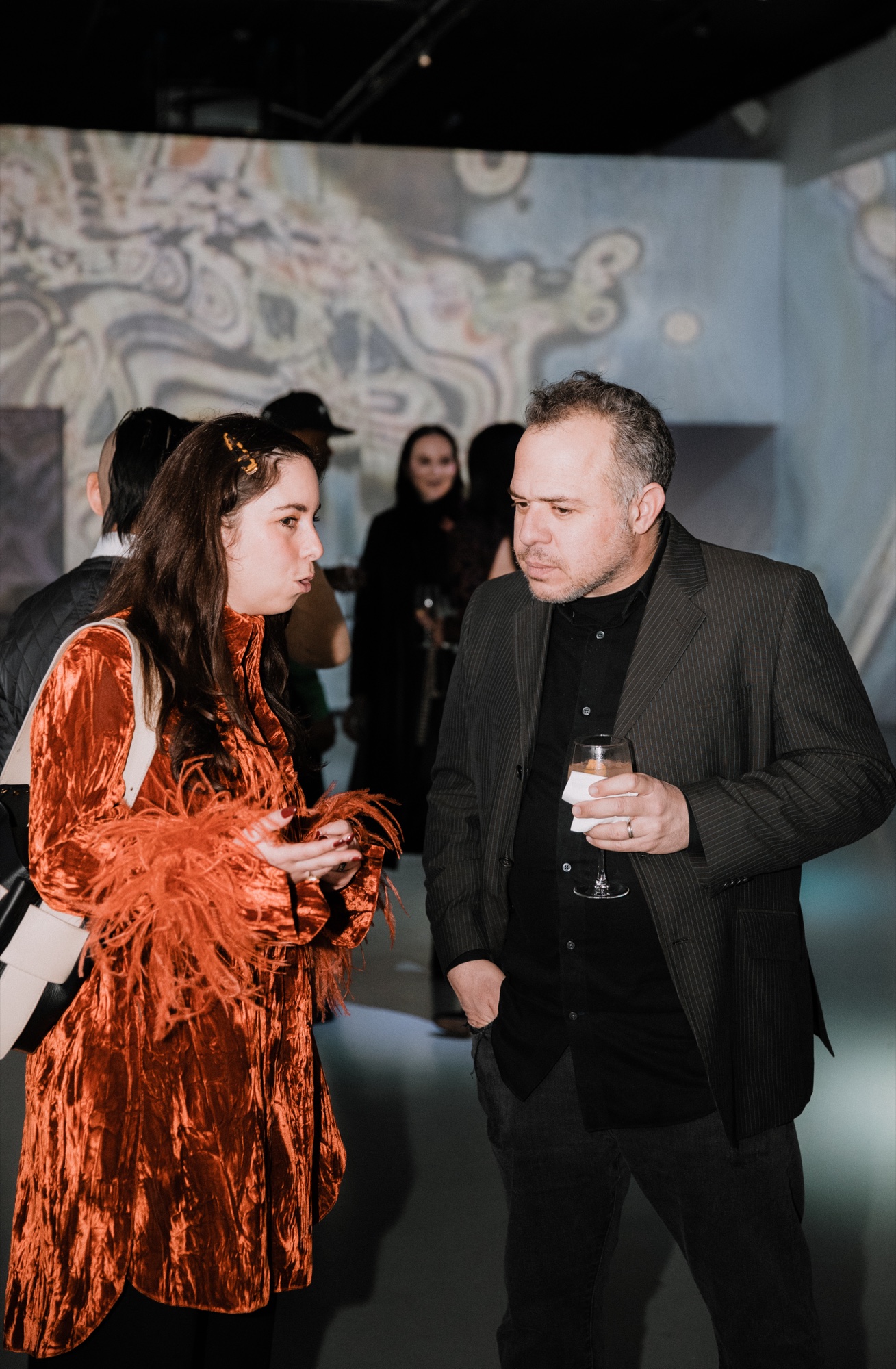 Woman in a rust velvet coat with feather trim talks with a man holding a drink under marbled projections