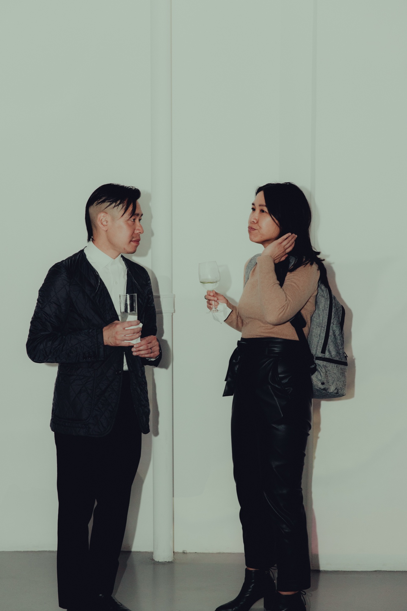 Two guests holding drinks talk in front of a plain white wall, one wearing a grey backpack