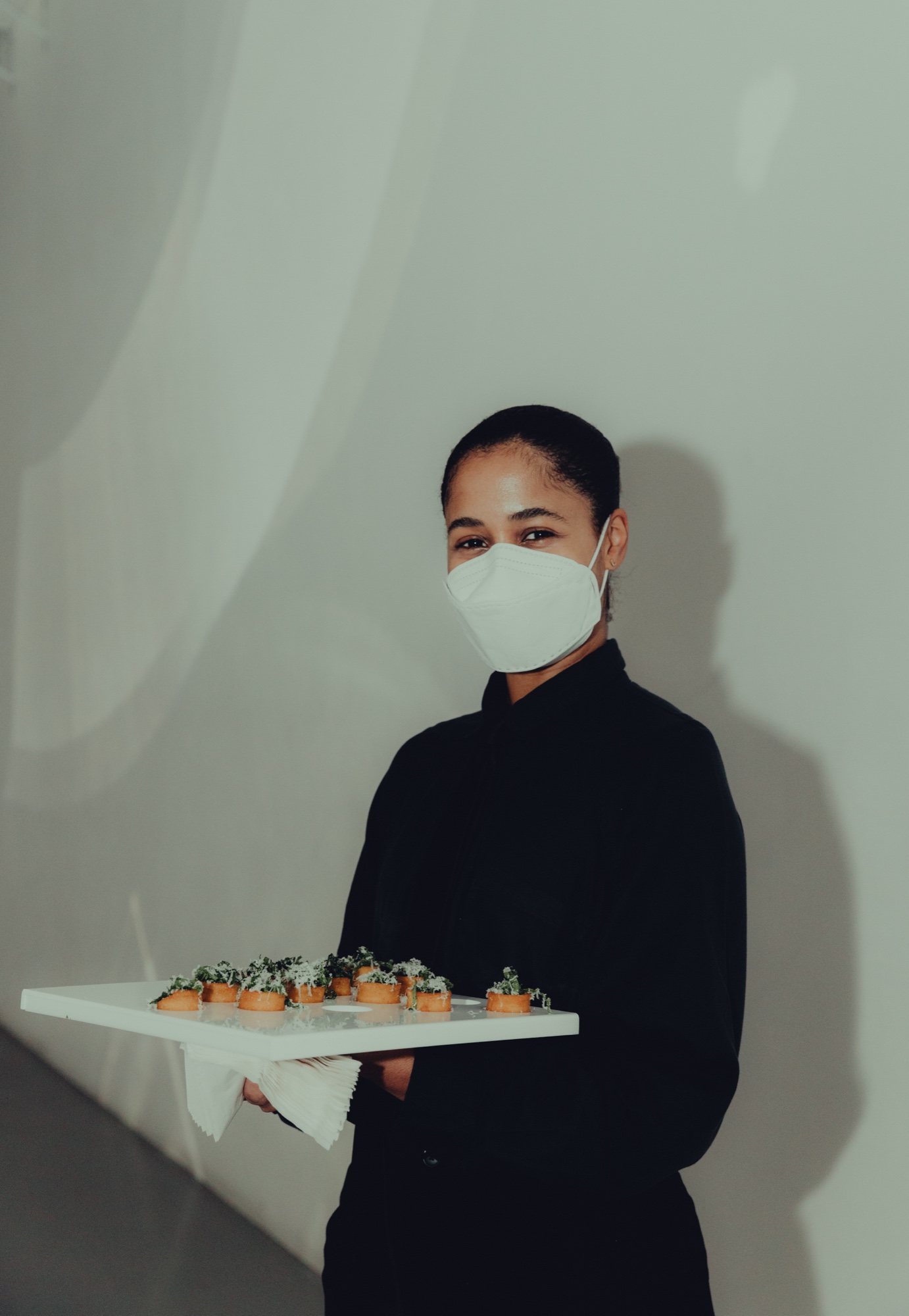 Server in a white mask and black shirt holds a tray of small tartlets against a pale grey wall