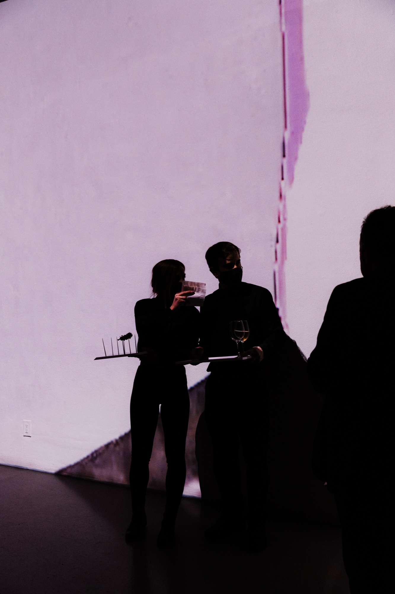 Two silhouetted servers with trays and a wine glass in front of a pale pink projected wall