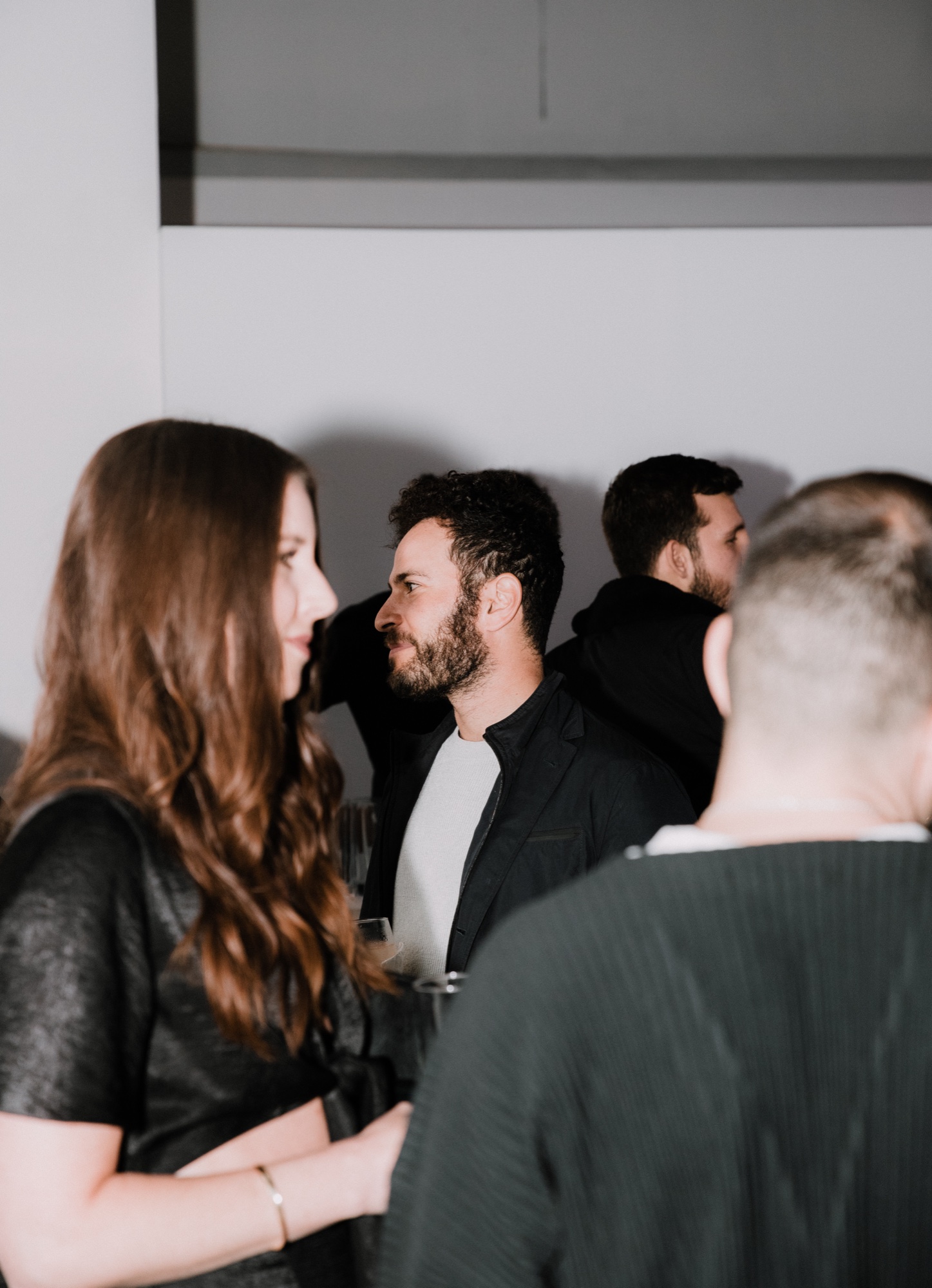 Guests in dark clothing gather close in a pale room, a bearded man in a black jacket in profile