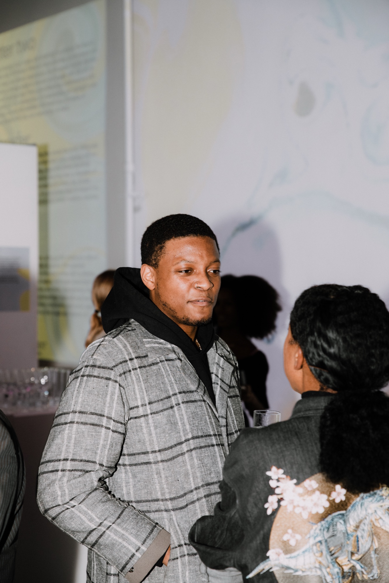 Man in a grey plaid coat and black hoodie talks with a guest before a wall of pale projected text