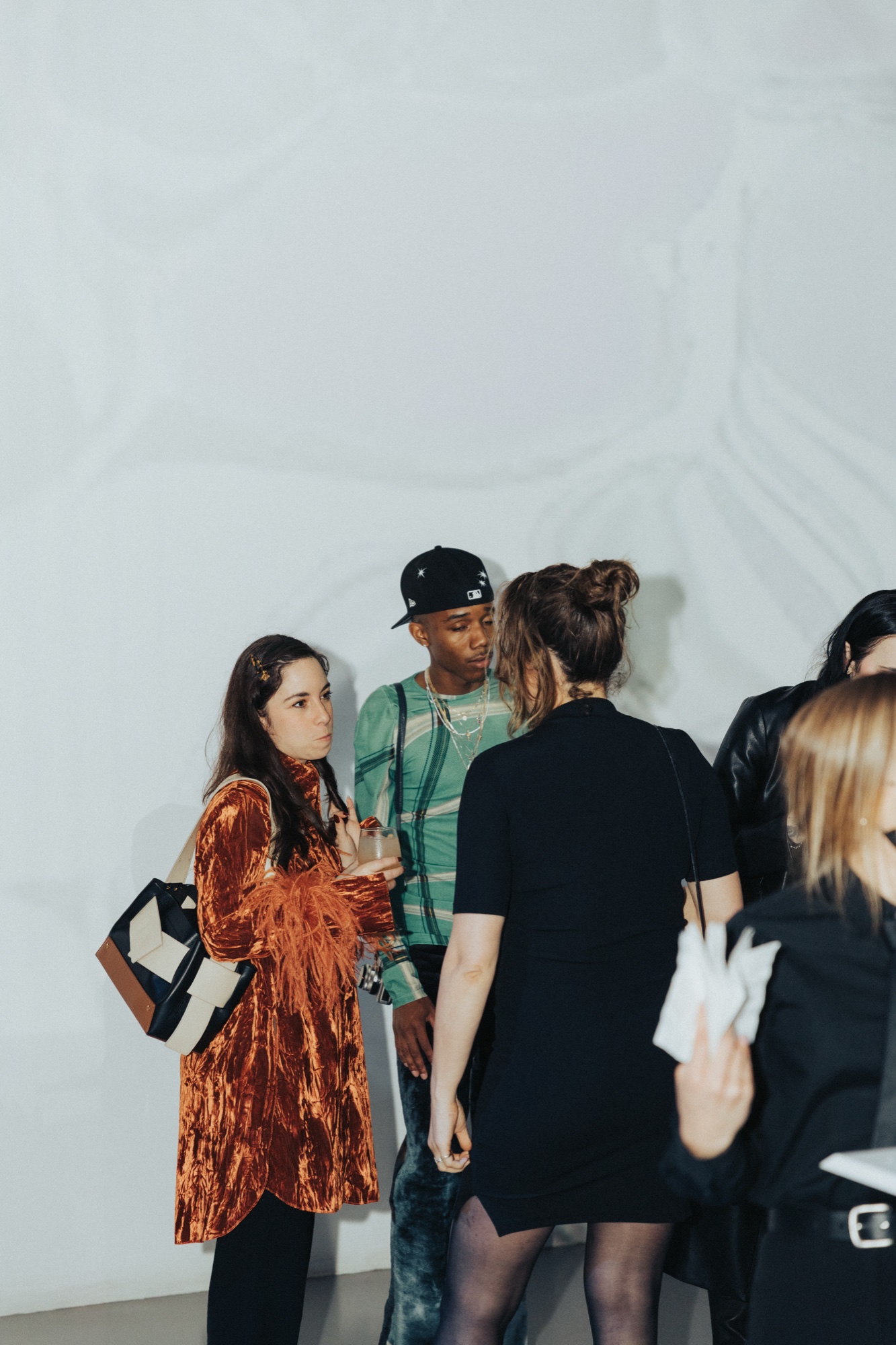 Guests in an orange velvet coat, green top and black dress chat before a white wall with rippling light