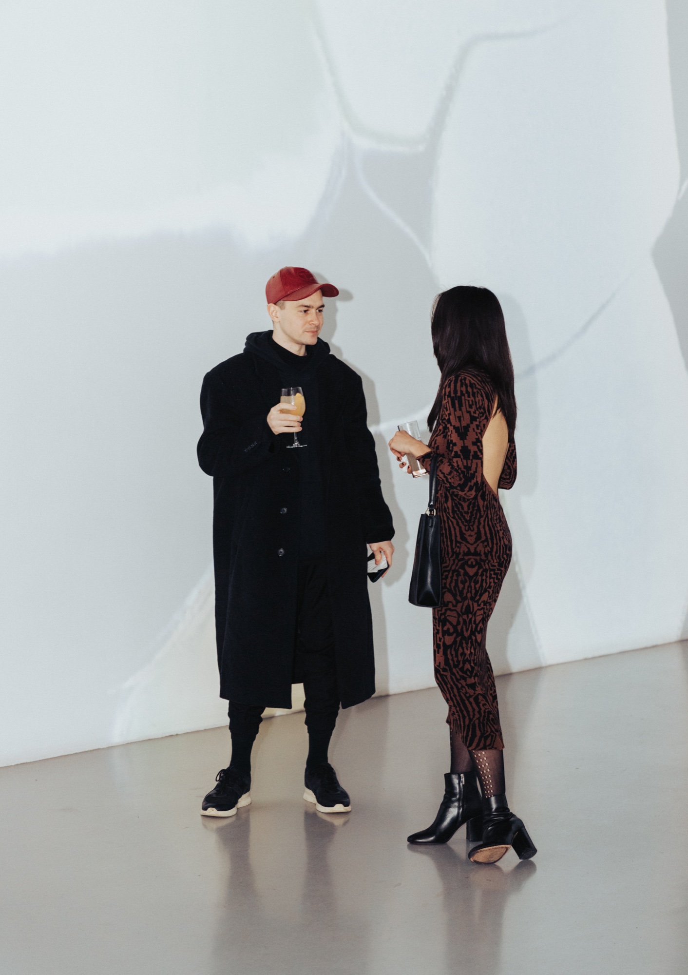 Man in a red cap and long black coat with a drink talks to a woman in a patterned dress