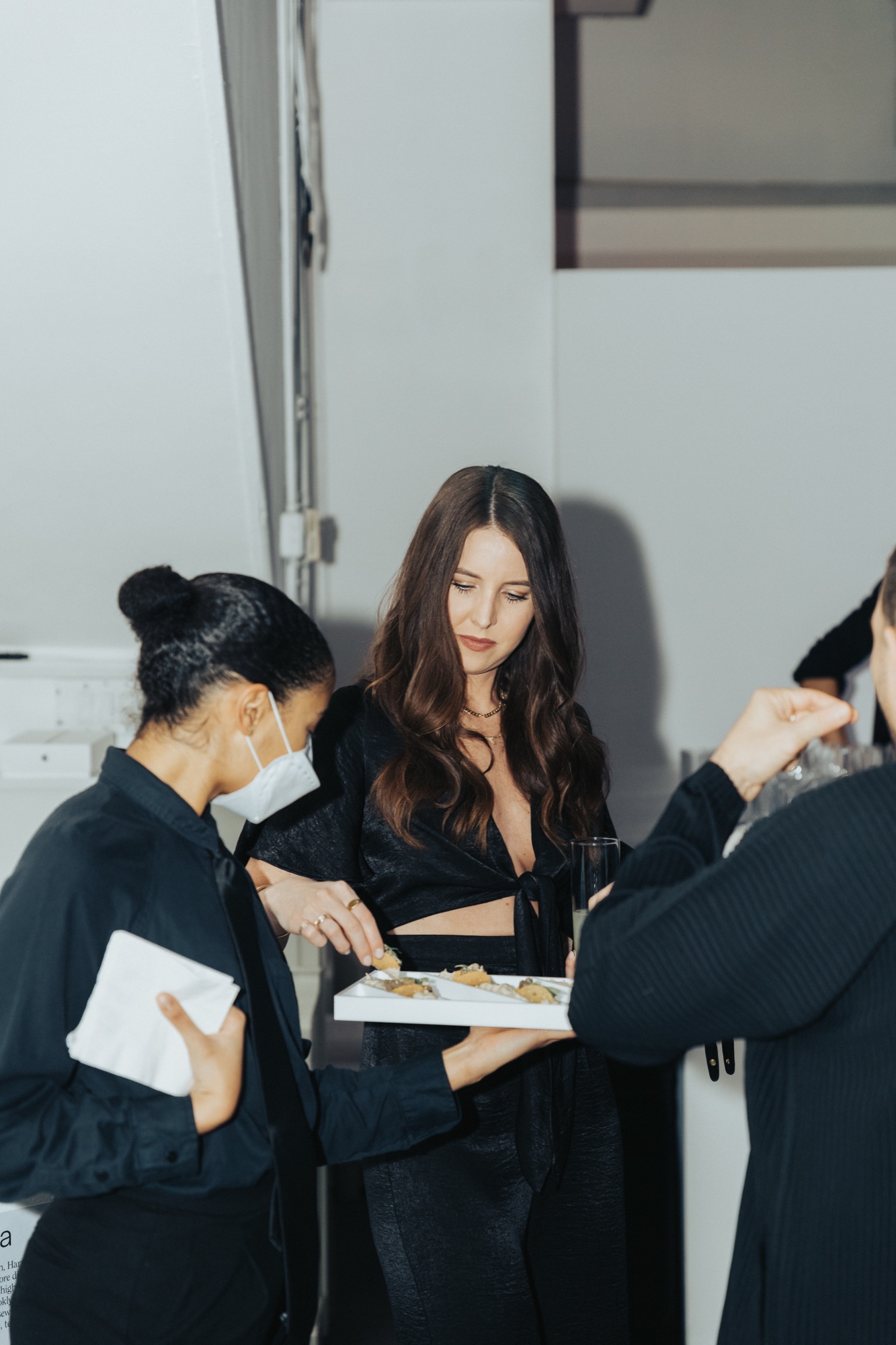 Woman in a white mask takes a canapé from a tray held by a long haired guest in black