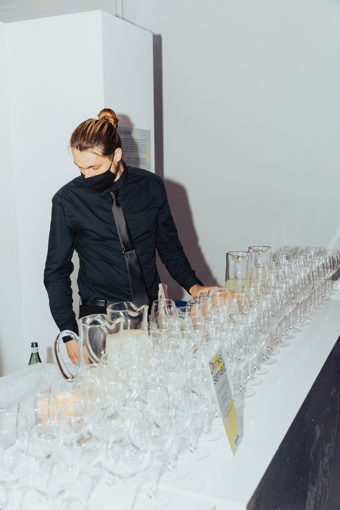 Bartender in a black shirt and mask fills glasses among rows of wine glasses and pitchers on a white counter