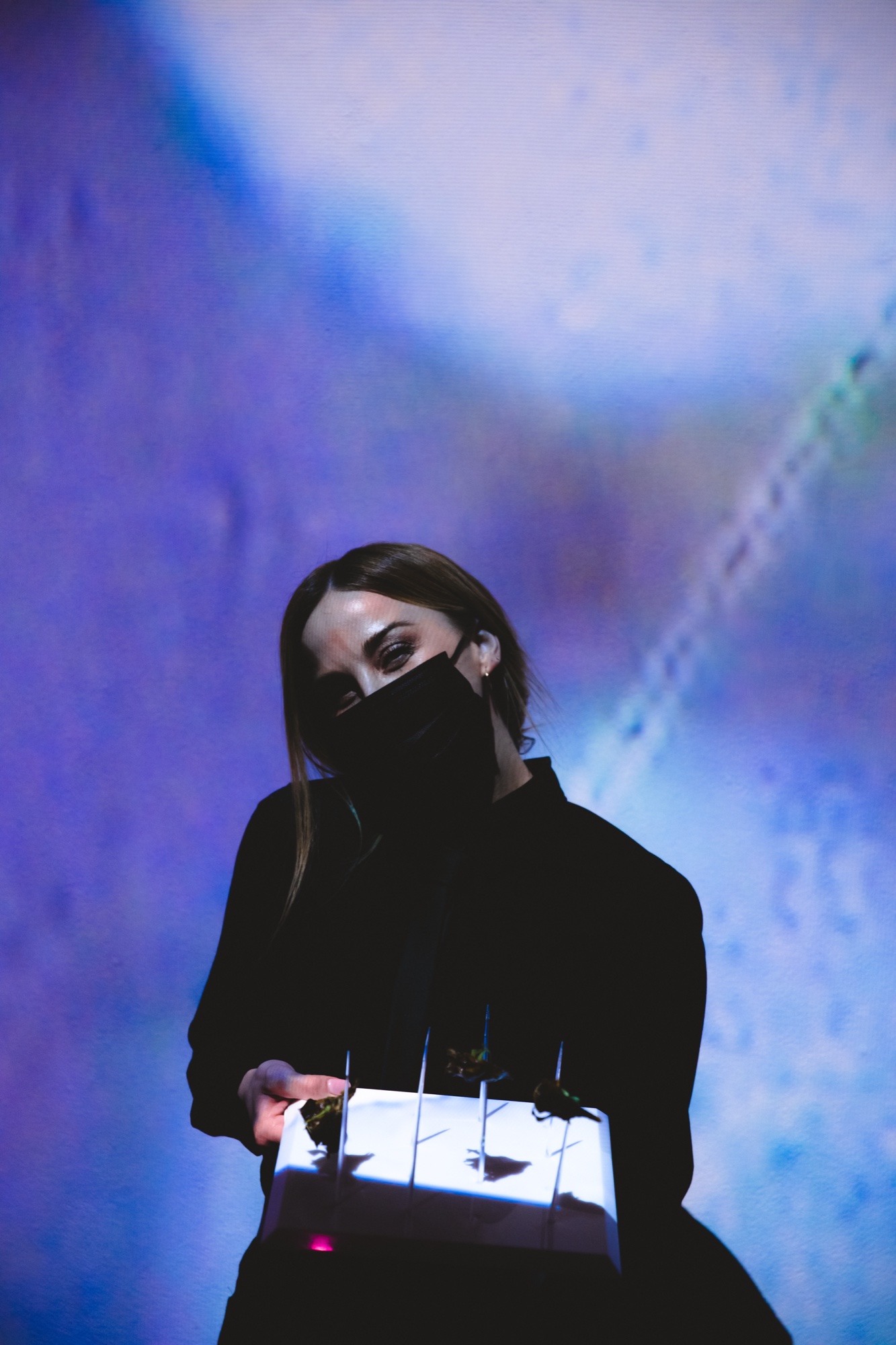 Woman in a black face mask holds a lit white tray with small sprouts against a blue projected wall