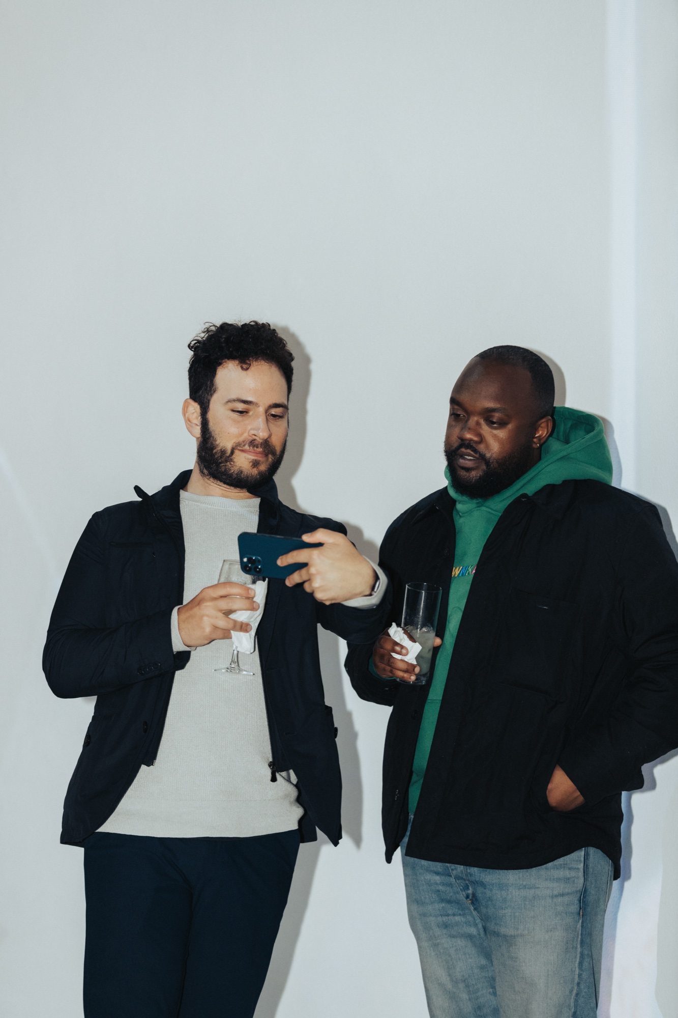 Two men lean on a white wall holding drinks, one showing the other a phone, one in a green hoodie