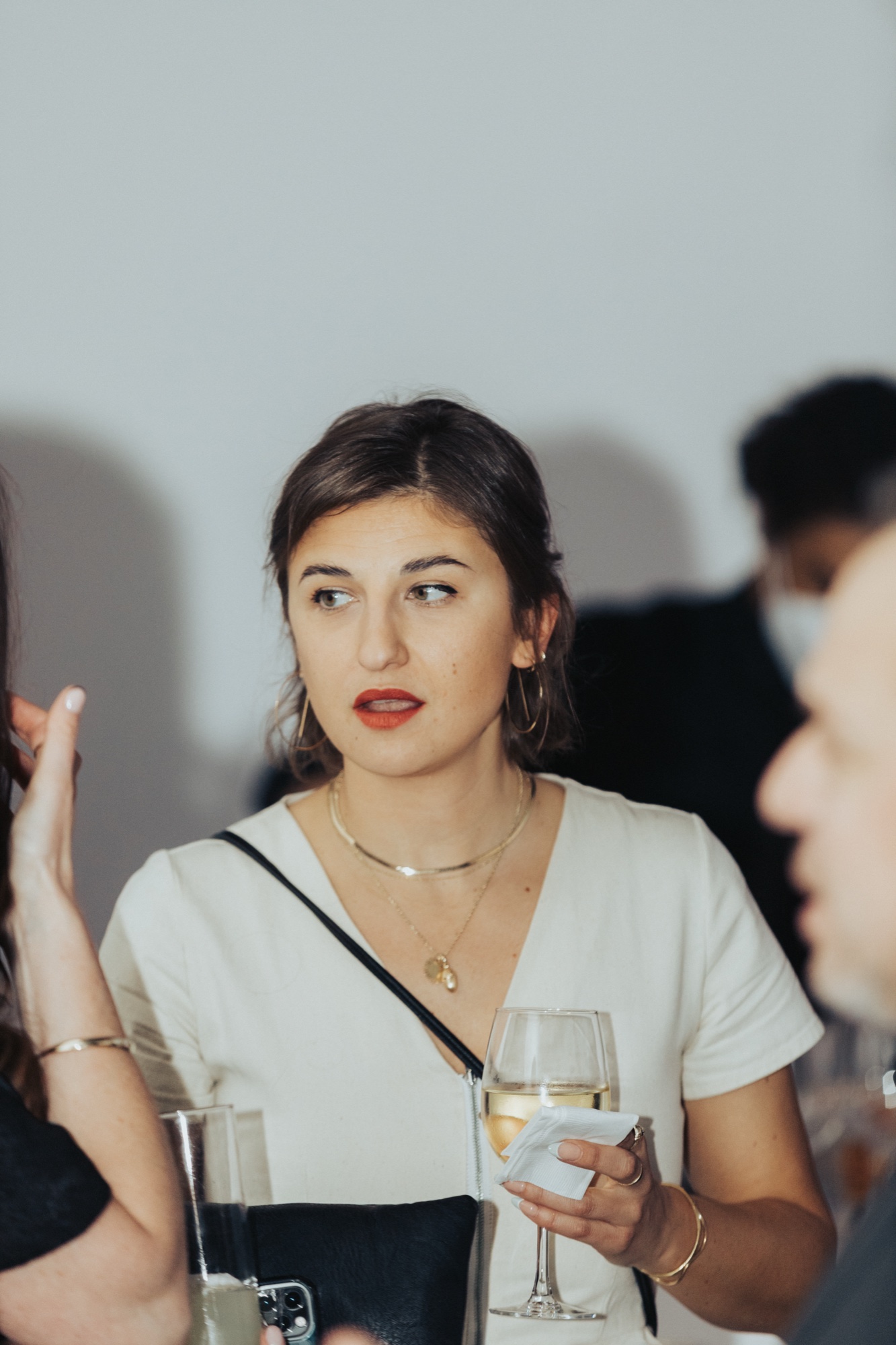 Woman in a white top with red lipstick holds a glass of white wine and glances sideways