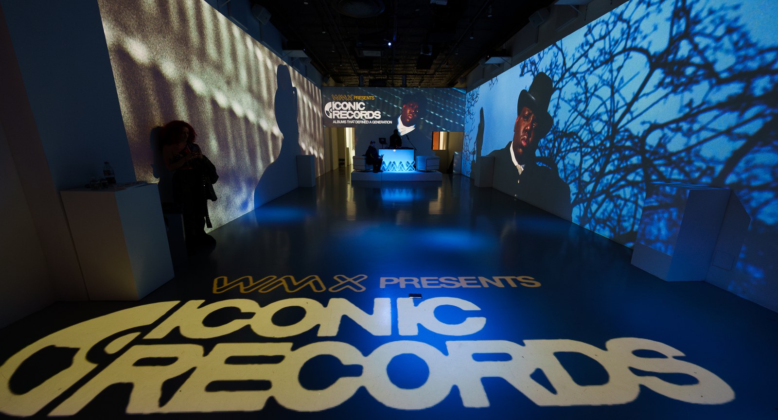 WMX Presents lettering projected in yellow on a dark floor, portrait footage on the blue lit walls ahead