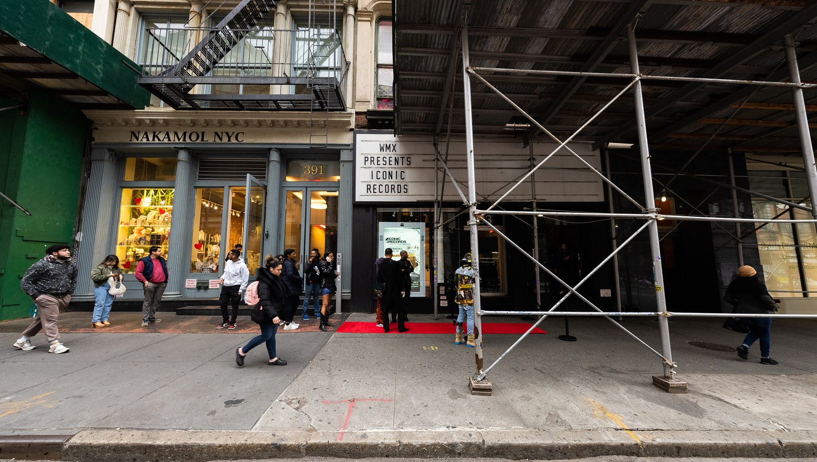 Pedestrians pass a red carpet entrance under a WMX Presents marquee on a scaffolded street