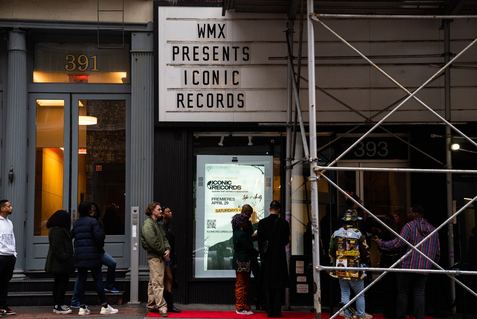 Guests queue on a red carpet below a WMX Presents Iconic Records marquee and a lit poster window