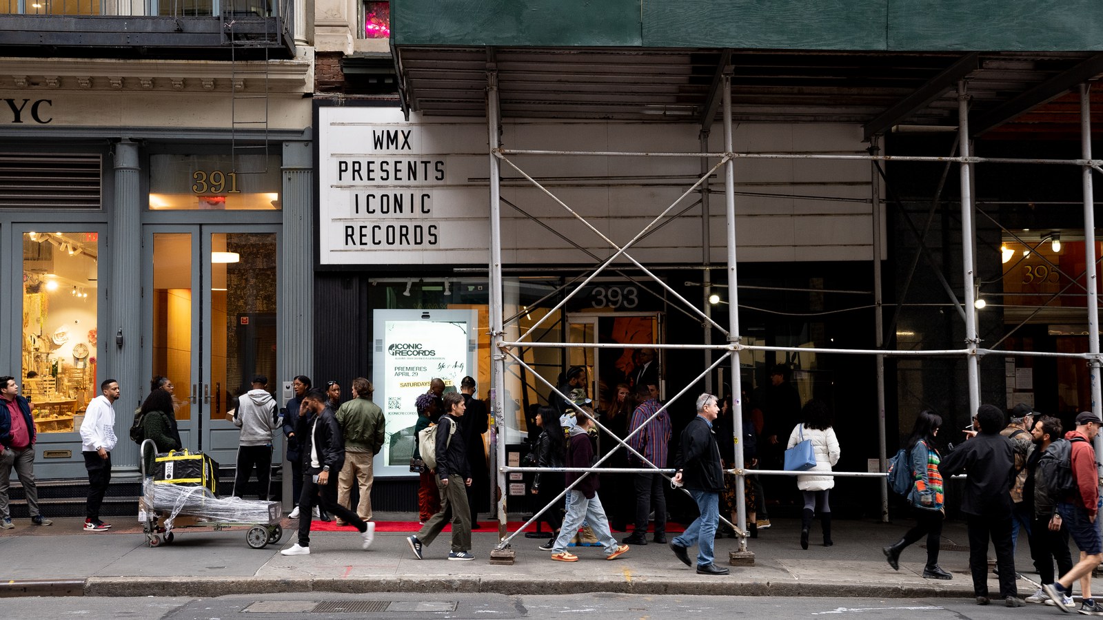 Crowd walks past a WMX Presents marquee and red carpet beneath scaffolding, one man pushing a cart