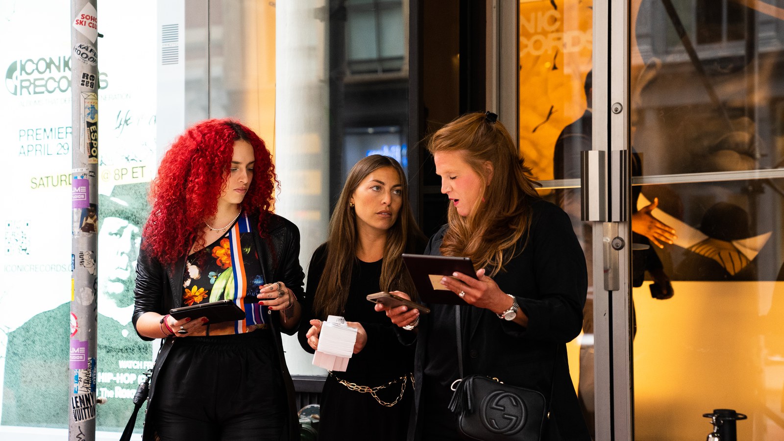 Three women check tablets and a phone at a glass entrance, one with bright red curls