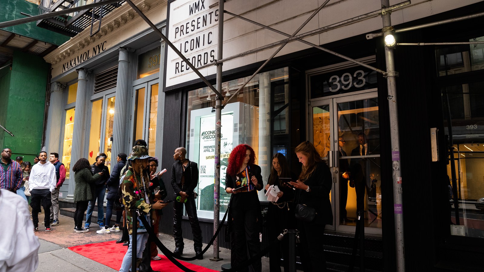 Woman with red curls and other guests wait at a stanchion rope on a red carpet beneath scaffolding