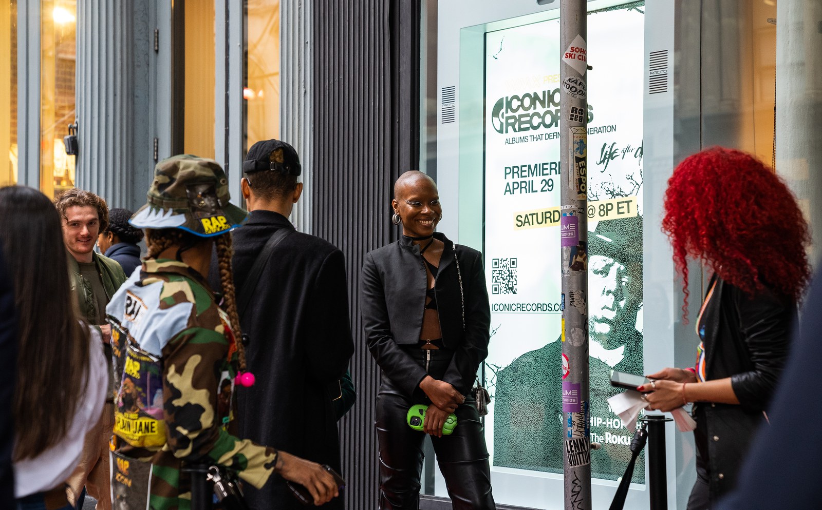 Guests line up outside a window poster announcing a premiere on April 29, a woman with red hair at right