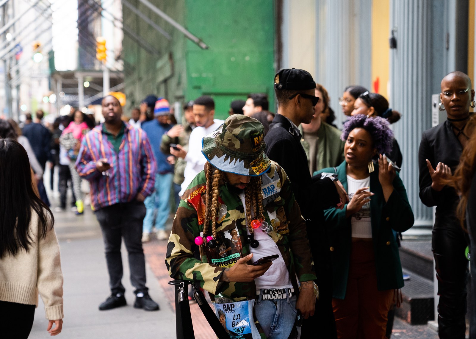 Person in a camouflage jacket with long braids checks a phone among a crowd waiting under sidewalk scaffolding