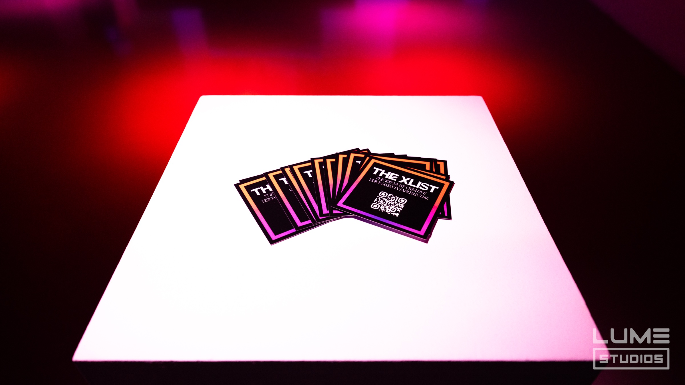 Fanned stack of The XLIST cards with QR codes on a white pedestal washed in red light