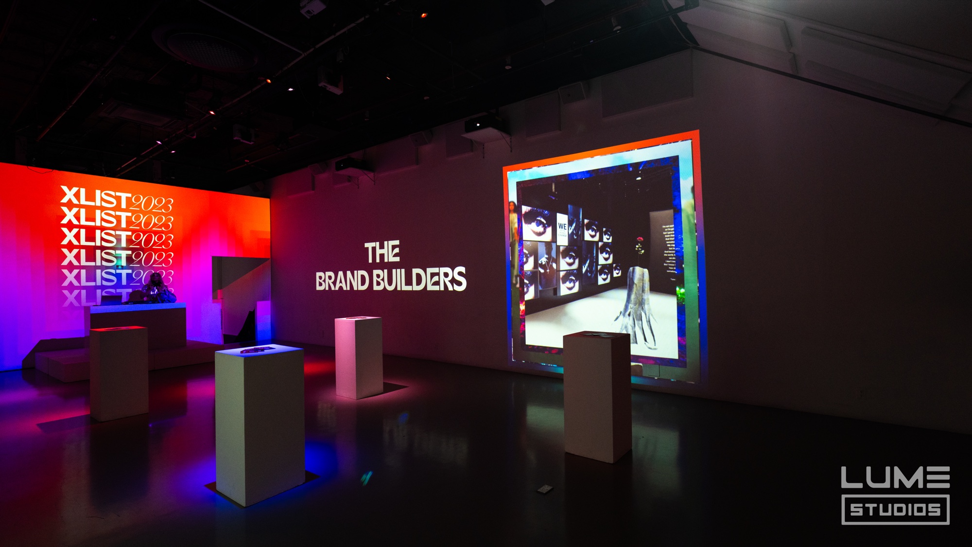 White pedestals face a dark wall reading The Brand Builders beside a colourful projected screen and a red XLIST panel