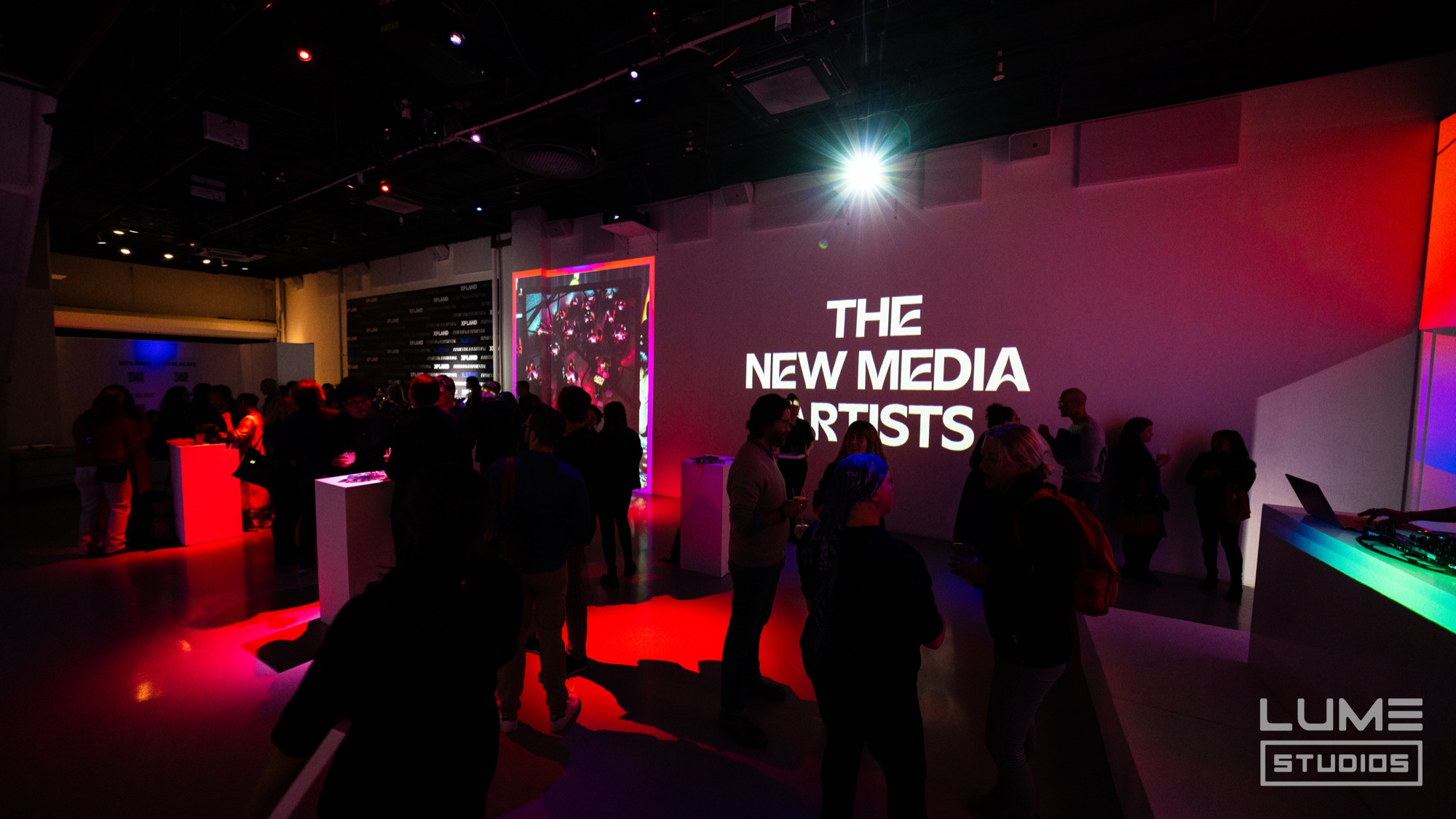 Guests mingle in a dark room lit red and purple before a projected sign reading The New Media Artists
