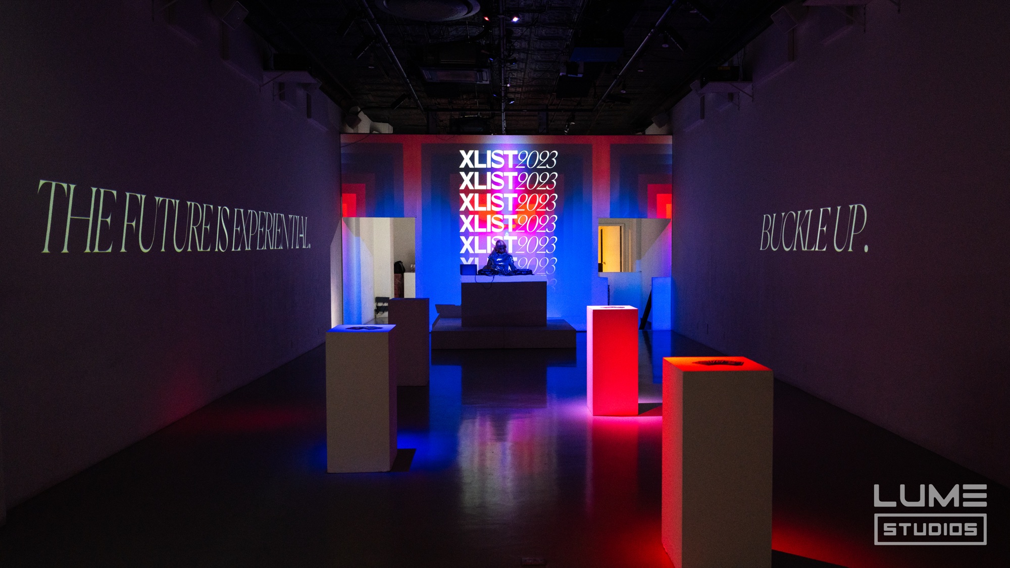 White plinths lit red and blue before an XList 2023 screen, walls reading The Future Is Experiential and Buckle Up