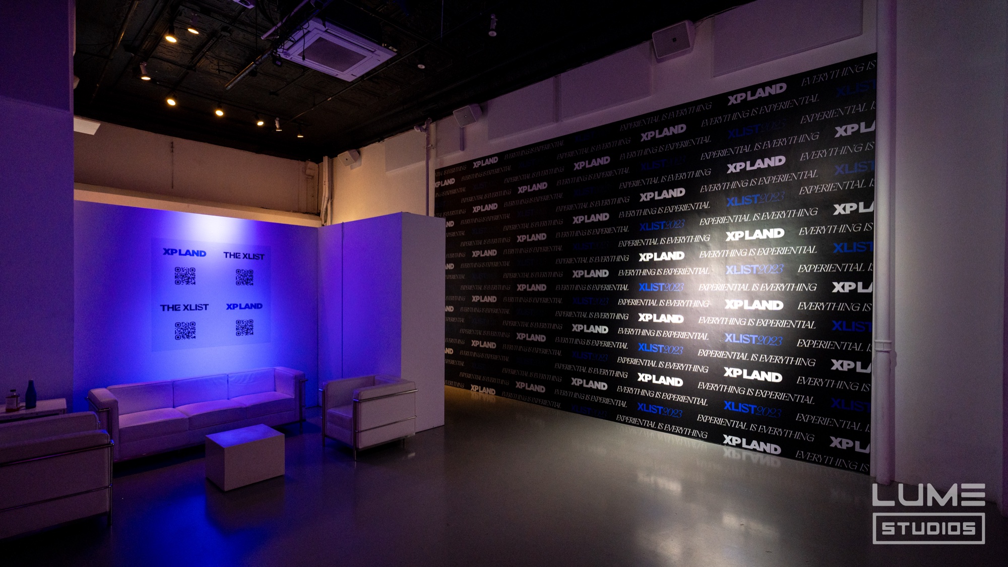 A white sofa lounge in purple light beside a black step and repeat wall covered in XP Land logos