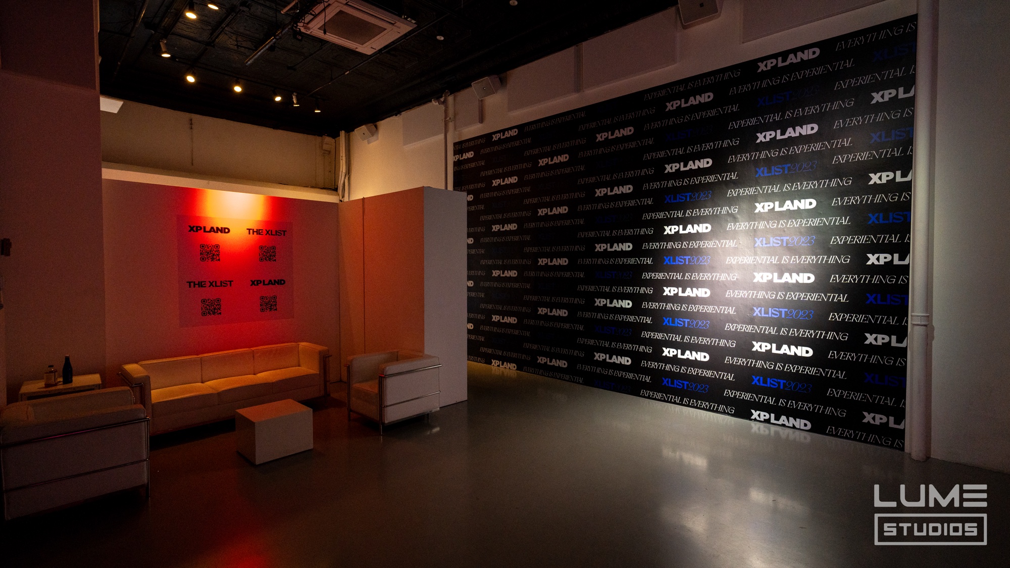 Lounge with white sofas beside a black XP Land logo wall under warm spotlights