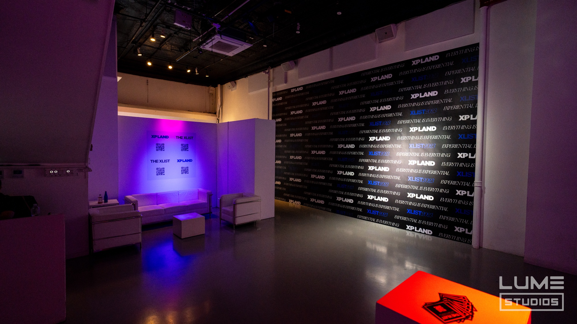 A black step and repeat wall printed with XP Land logos stands beside a white lounge set lit purple