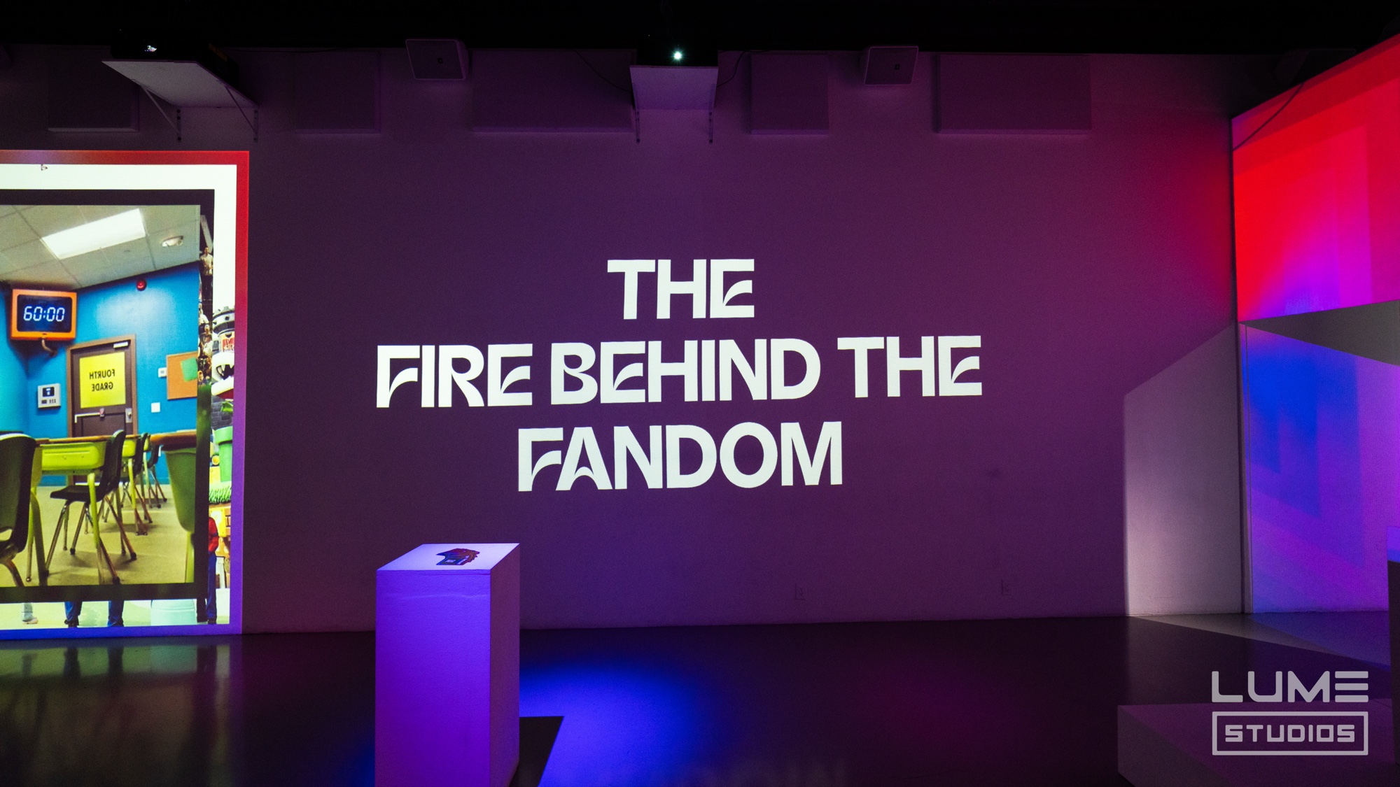 A dark stage wall projected with The Fire Behind the Fandom in white letters beside a screen showing a classroom