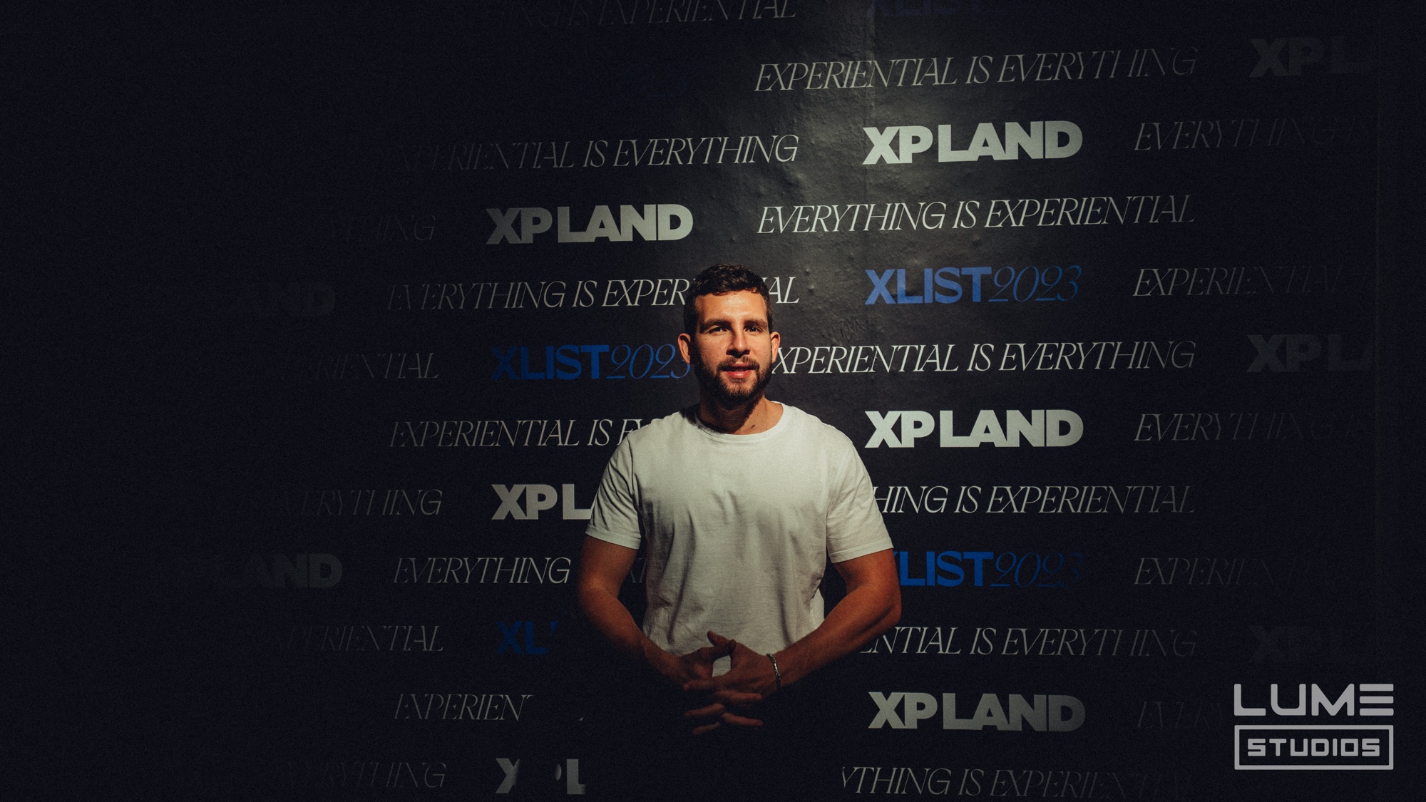 A man in a white shirt stands before a dark wall printed with XP Land and Experiential Is Everything