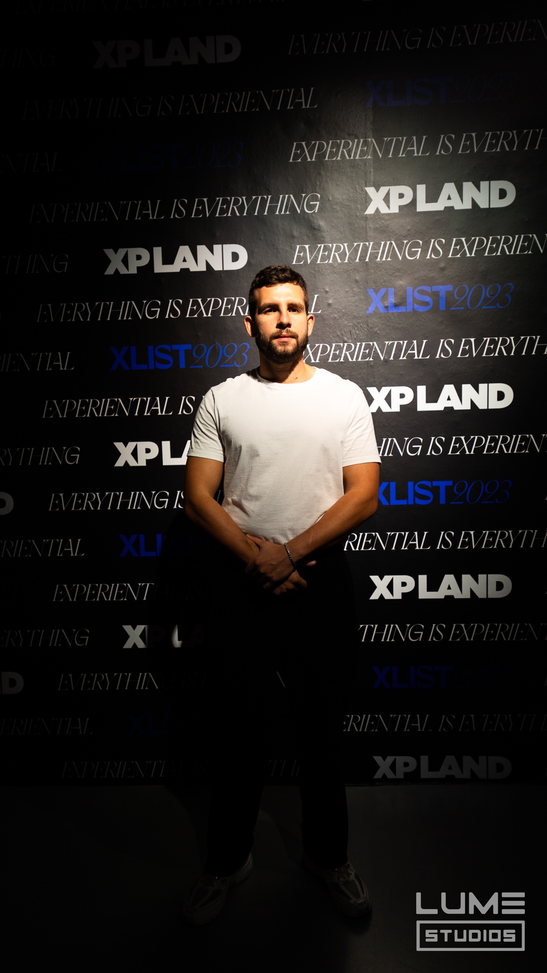 A man in a white tee stands spotlit before a black wall repeating XP Land and Experiential Is Everything